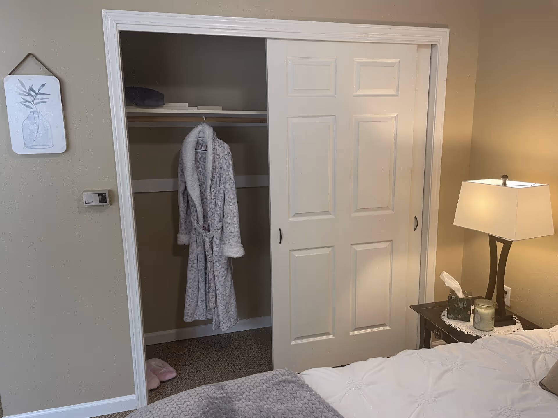 A bedroom corner featuring a partially open white sliding closet door with a hanging gray patterned robe inside. Next to the closet is a nightstand with a lit lamp, a tissue box, and a candle. The bed with a white comforter and gray knitted blanket is partially visible in the foreground. A small framed artwork of a plant in a vase hangs on the beige wall beside a thermostat.