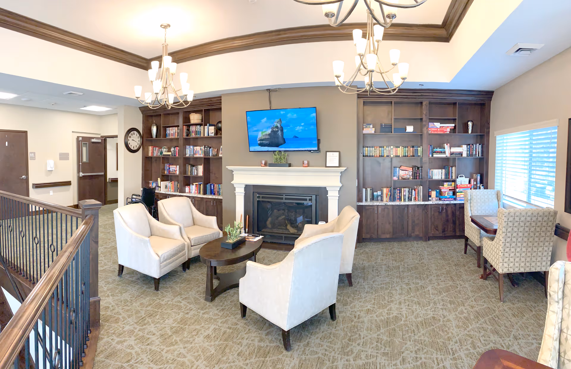 A cozy assisted living common area with four beige armchairs arranged around a small oval wooden coffee table. Behind the chairs is a fireplace with a flat-screen TV mounted above it. On either side of the fireplace are built-in dark wood bookshelves filled with books and games. To the right, there is a window with blinds and a small table with two patterned chairs. The room has beige walls, carpeted floor, and two chandeliers hanging from the ceiling.