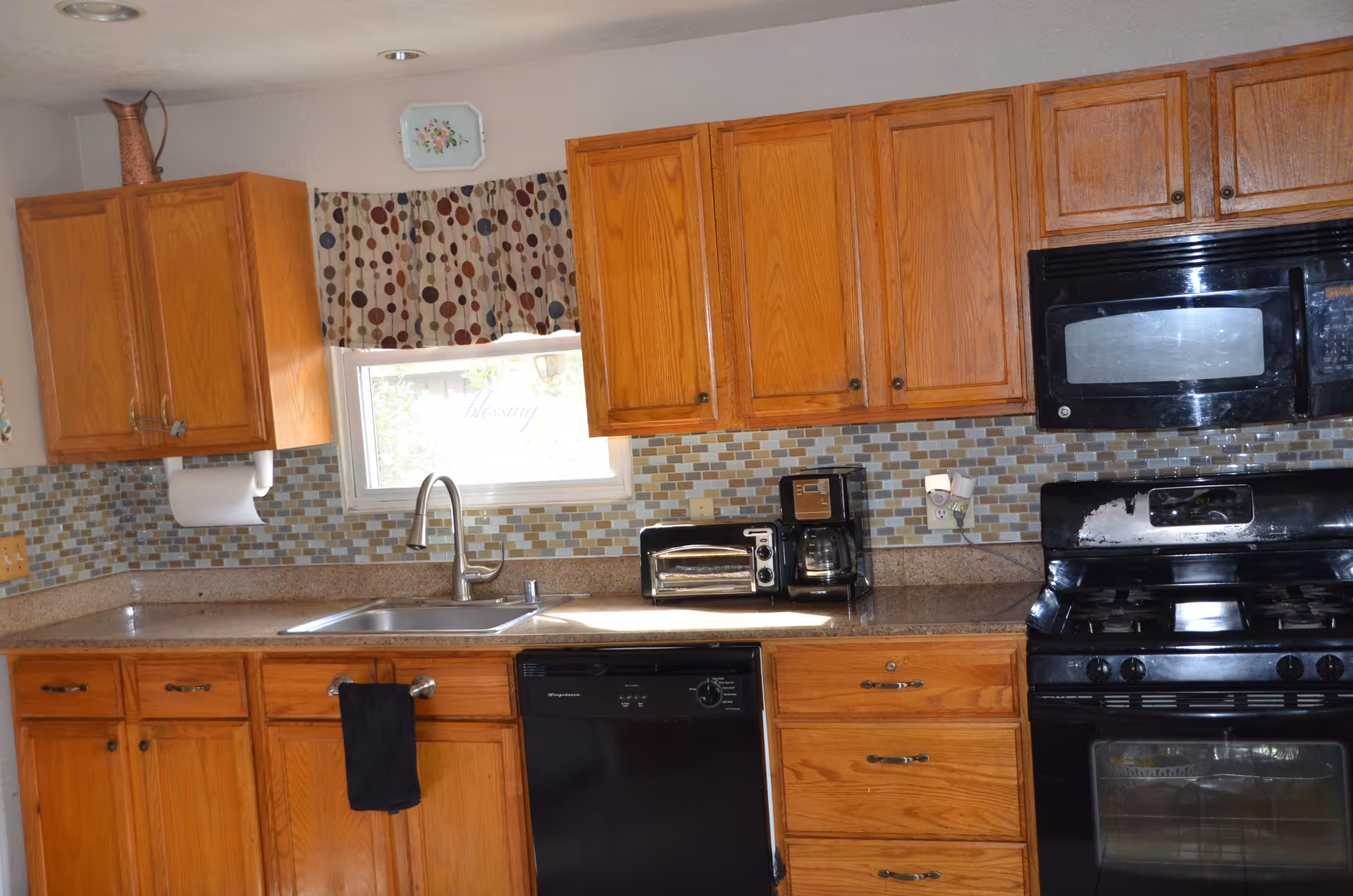 A kitchen with wooden cabinets, a stainless steel sink under a window with a patterned valance, a black dishwasher, a black stove with an oven, a black microwave above the stove, a toaster oven, and a coffee maker on the countertop. The backsplash is made of small square tiles in various shades of beige and brown.