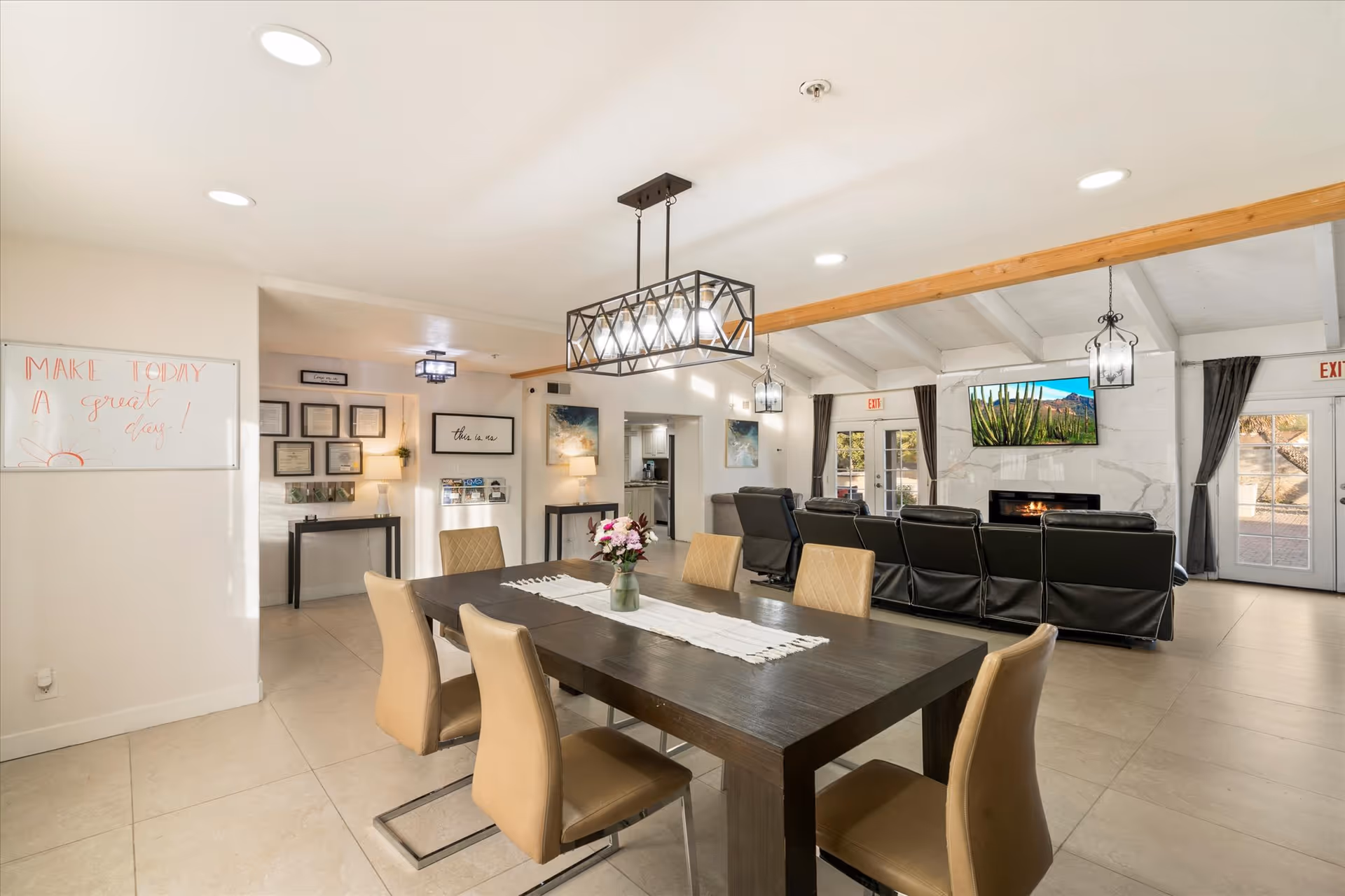 A bright and spacious common area in an assisted living facility featuring a dark wooden dining table with six beige chairs, a vase with flowers on the table, a modern rectangular chandelier above, and a seating area with black recliners facing a wall-mounted TV above a fireplace. The room has white walls, large windows with curtains, and a whiteboard on the left wall with the message 'MAKE TODAY a great day!' written on it.