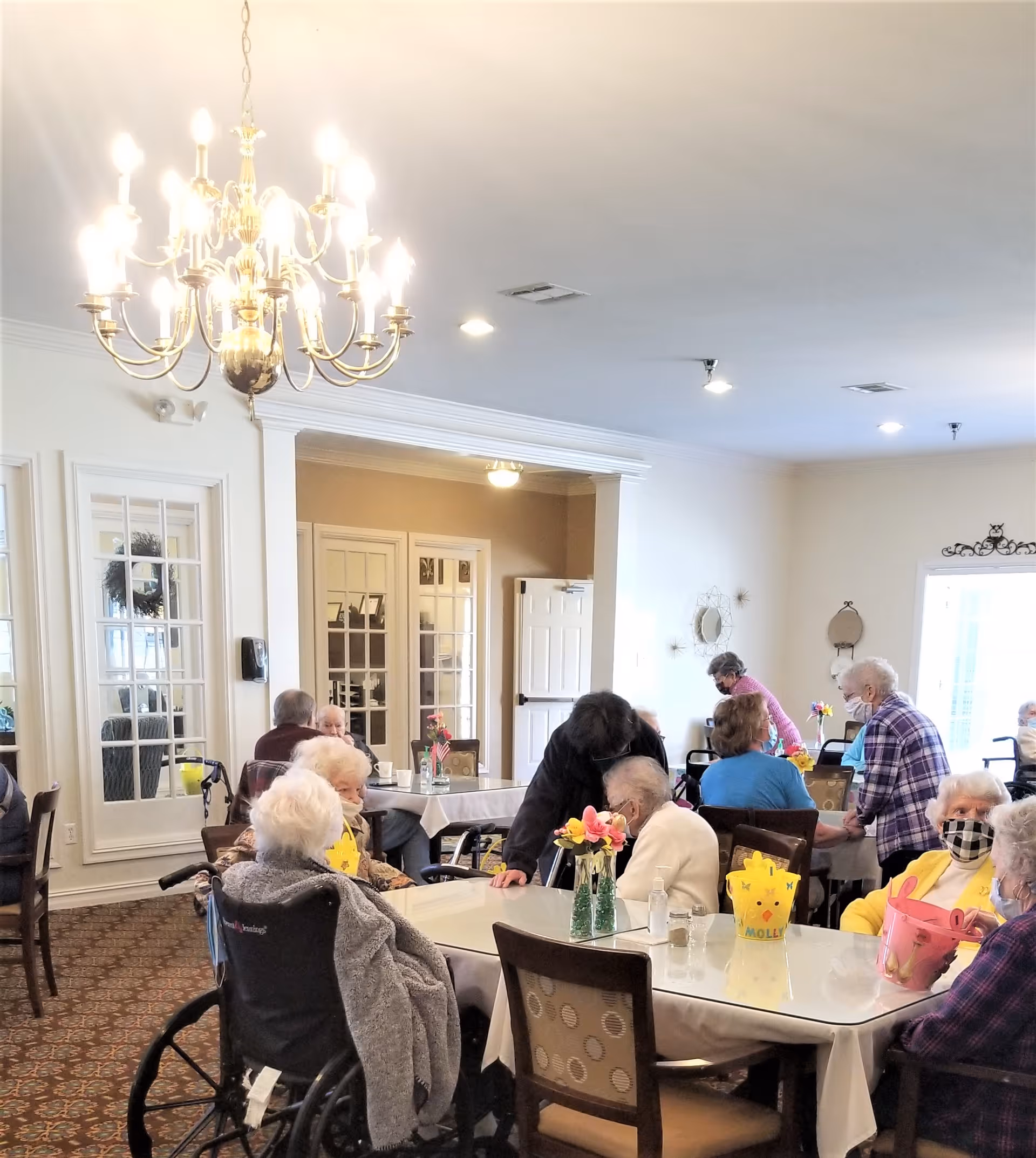 Elderly residents seated and chatting around tables in a bright communal dining room with a chandelier overhead.