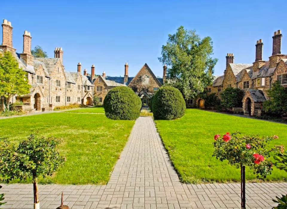 A well-maintained courtyard with a paved walkway leading through green lawns and neatly trimmed bushes, surrounded by stone buildings with multiple chimneys under a clear blue sky.
