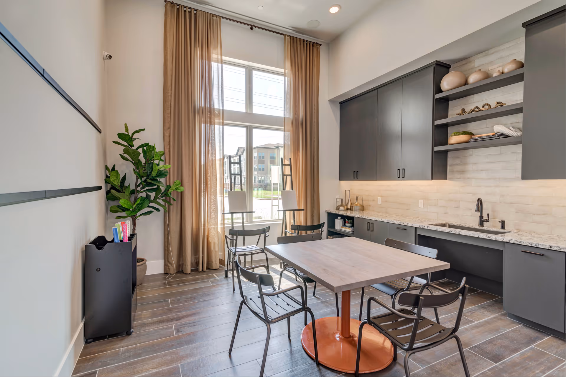 Bright communal dining area with a central table and chairs, dark cabinetry and sink along one wall, large window with floor-length curtains, and a potted plant.