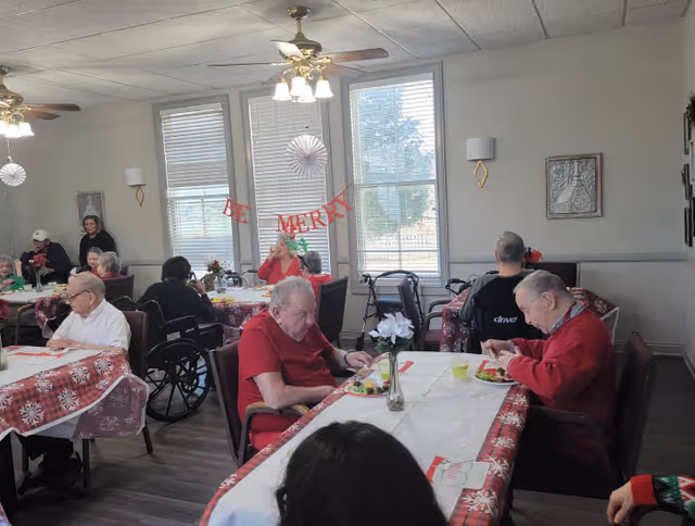 A dining room in a senior living facility with elderly residents seated at tables covered with festive red and white tablecloths. The room has large windows with blinds, ceiling fans with lights, and holiday decorations including a 'Be Merry' banner. Some residents are eating while others are engaged in conversation.