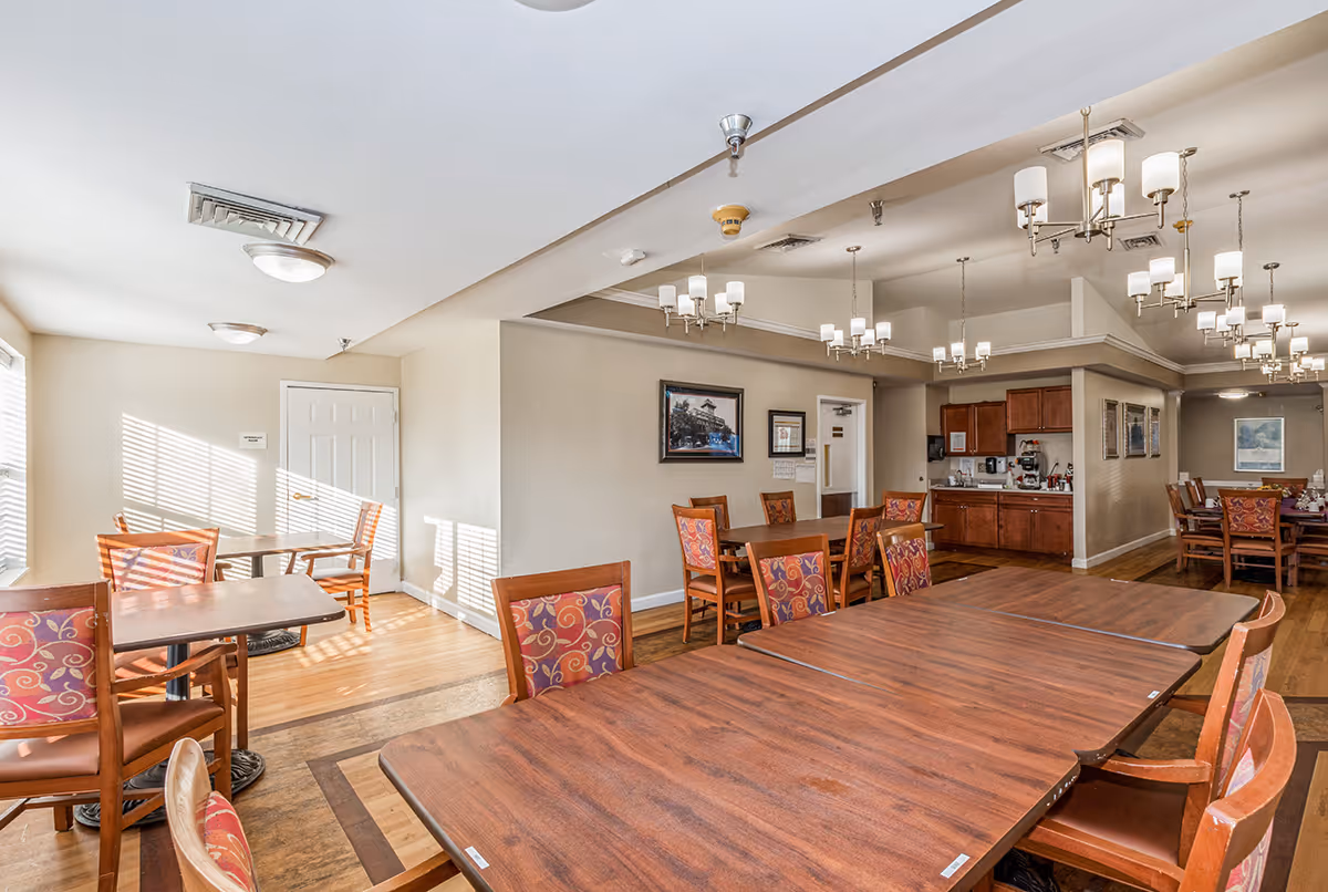 A bright dining room with multiple wooden tables and chairs featuring patterned cushions. The room has hardwood floors, beige walls, and several chandeliers hanging from the ceiling. There is a small kitchenette area with cabinets and a coffee machine in the background, and framed pictures on the walls.