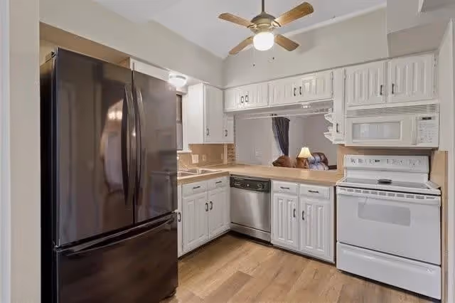 A modern kitchen with white cabinets, a black refrigerator, a white oven with a microwave above it, a dishwasher, and a ceiling fan with a light. The kitchen has wooden flooring and a pass-through window looking into a living area with a lamp and curtains.
