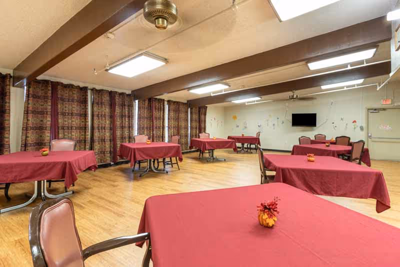 A spacious dining room with multiple tables covered in red tablecloths, each table decorated with a small centerpiece. The room has wooden flooring, patterned curtains covering the windows, ceiling fans, and fluorescent lighting. A wall-mounted TV is visible on the far wall.