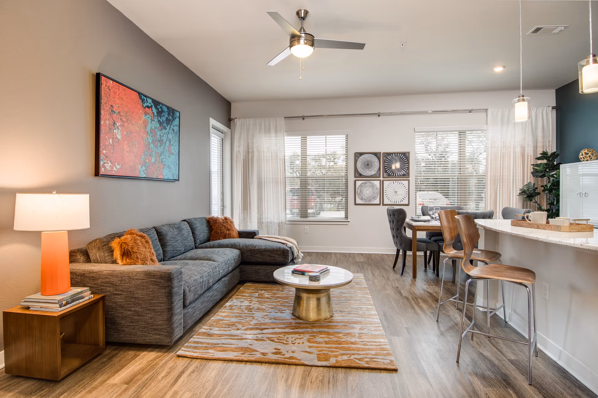A modern living room with a gray sectional sofa adorned with two orange throw pillows, a wooden side table with a lamp and books, a round coffee table on a patterned rug, and a dining area with a table and chairs near large windows with white curtains. The room has wood flooring, a ceiling fan, and contemporary wall art.