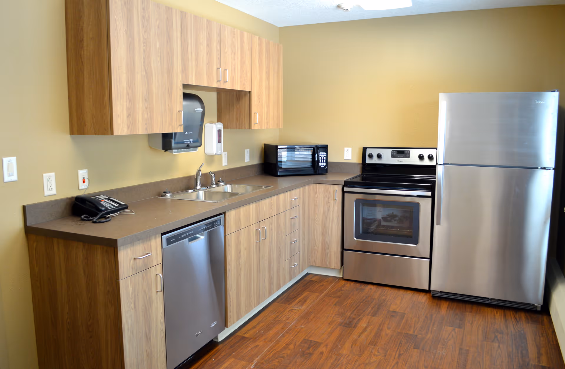 A clean kitchen area with wood cabinets, a stainless steel refrigerator, oven, dishwasher, a microwave on the countertop, a double sink, a wall-mounted paper towel dispenser, and a telephone on the counter. The floor has a wood finish and the walls are painted beige.