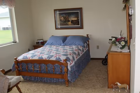 A bedroom with a wooden bed frame featuring a quilted bedspread in a star pattern and two blue pillows. There is a window on the left side letting in natural light, a wooden dresser with a floral arrangement on the right, and a framed painting hanging on the wall above the bed. The room has beige carpet and light-colored walls.