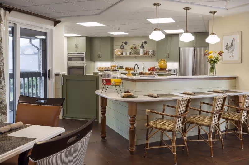A bright kitchen area with green cabinets, stainless steel appliances, and a large island with a white countertop. Four wicker bar stools are lined up at the island, which is set with placemats and napkins. Pendant lights hang above the island, and a vase with flowers is on the counter. To the left, there is a dining table with chairs and a sliding glass door leading outside.