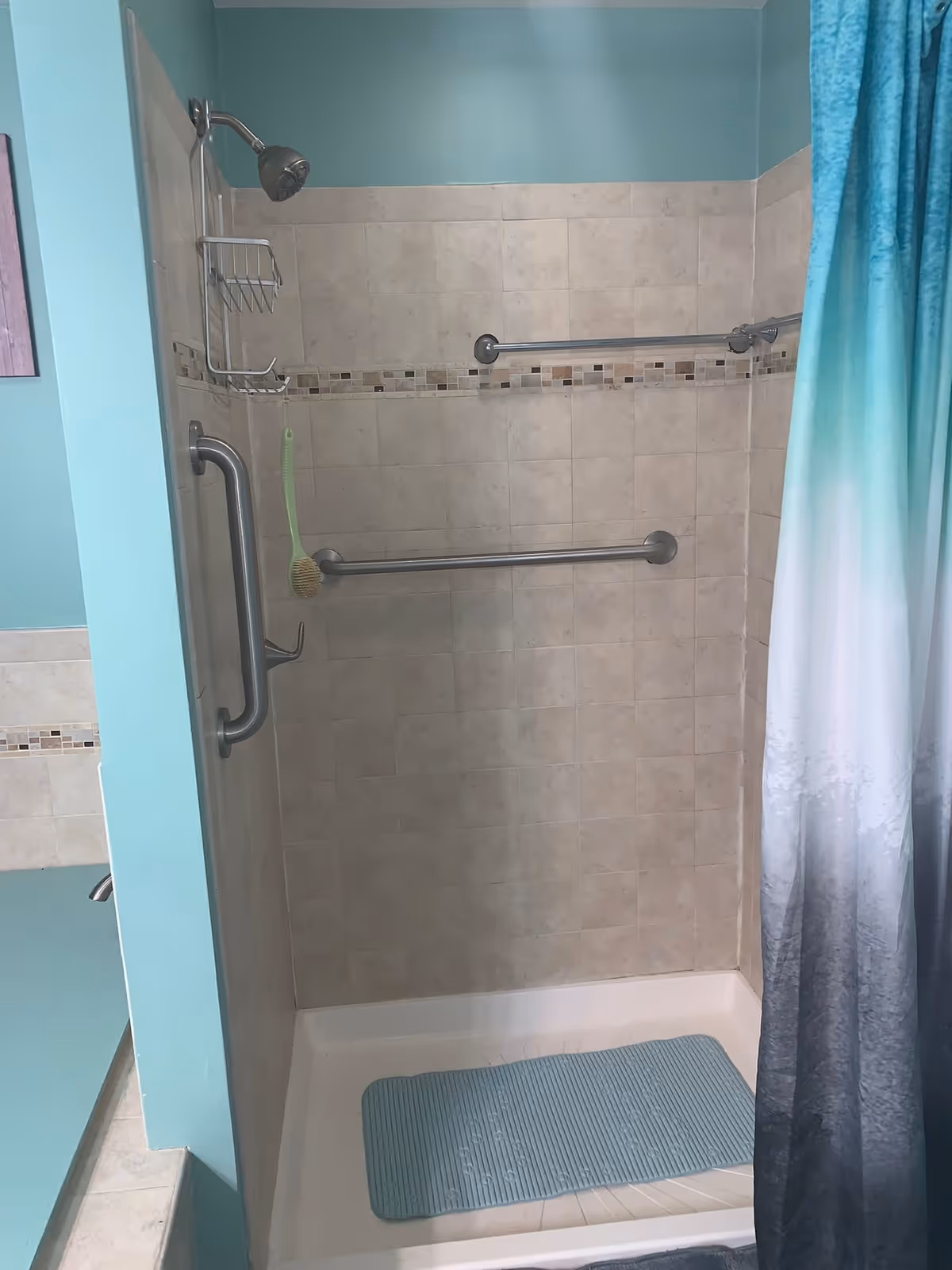 A tiled shower area with beige tiles and a decorative tile border. The shower has a silver showerhead, two silver grab bars, a hanging brush, and a blue non-slip mat on the shower floor. A blue and white gradient shower curtain is partially visible on the right side. The surrounding walls are painted light blue.