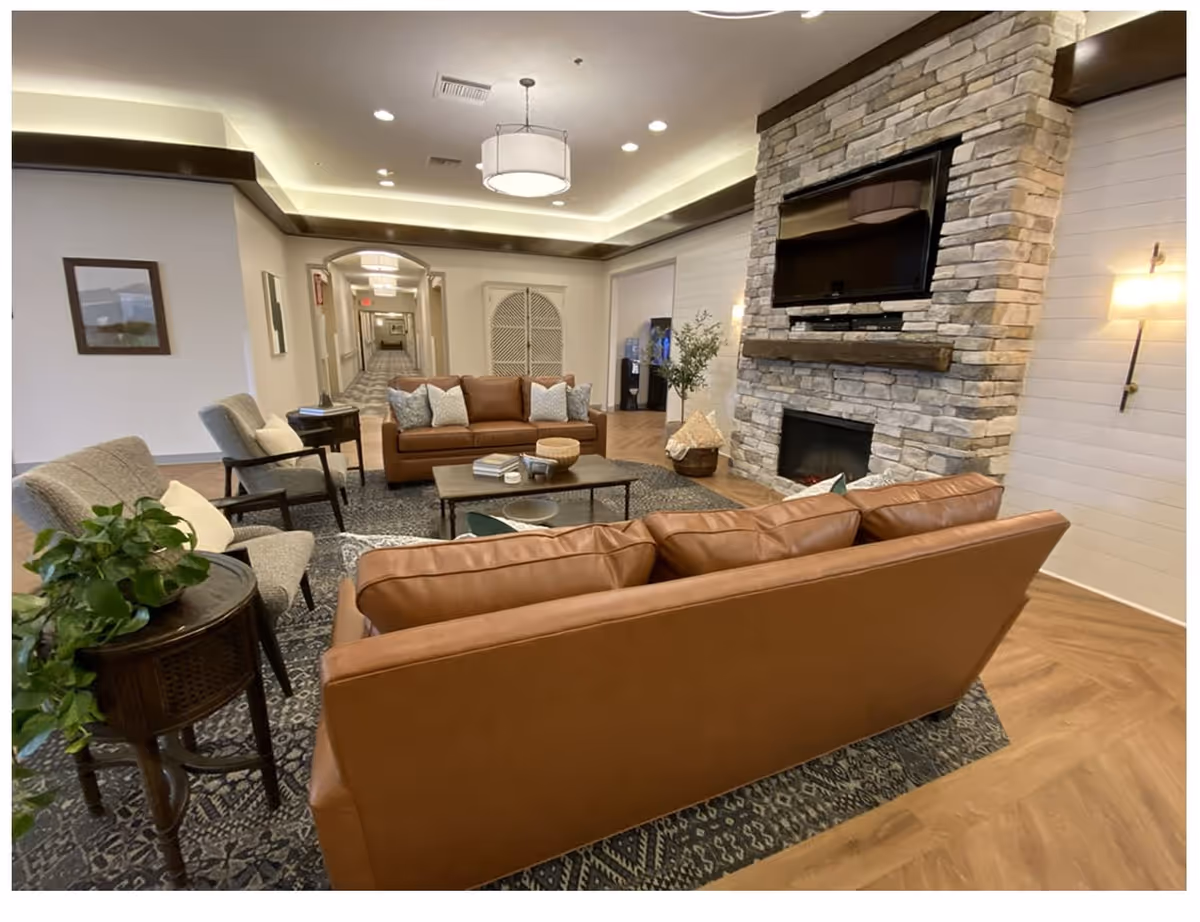 A cozy communal living room with brown leather sofas and armchairs arranged around a coffee table in front of a stone fireplace with a mounted TV.
