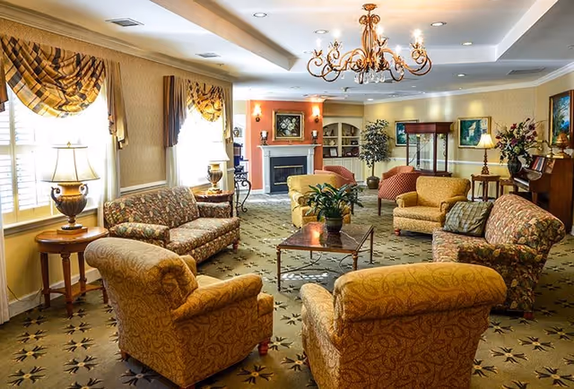 Well-lit communal living room with upholstered armchairs and sofas arranged around a central coffee table, a chandelier overhead, and a fireplace in the background.