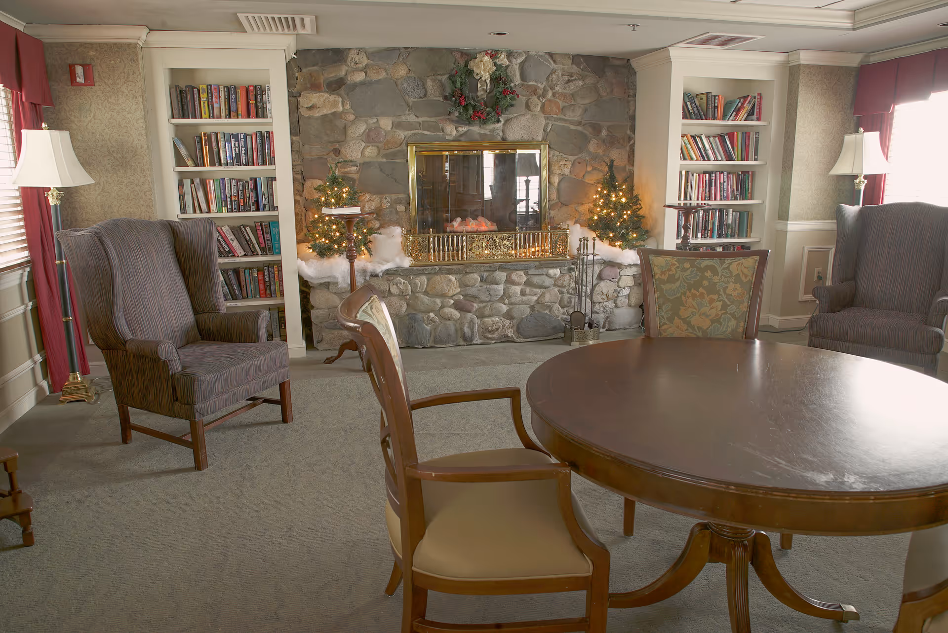 A cozy common room with a round wooden table, upholstered armchairs, bookshelves and a stone fireplace decorated with small lit trees.