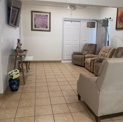 Tile-floored common room with beige reclining sofas, a mounted TV, side tables, framed artwork and a floor lamp.