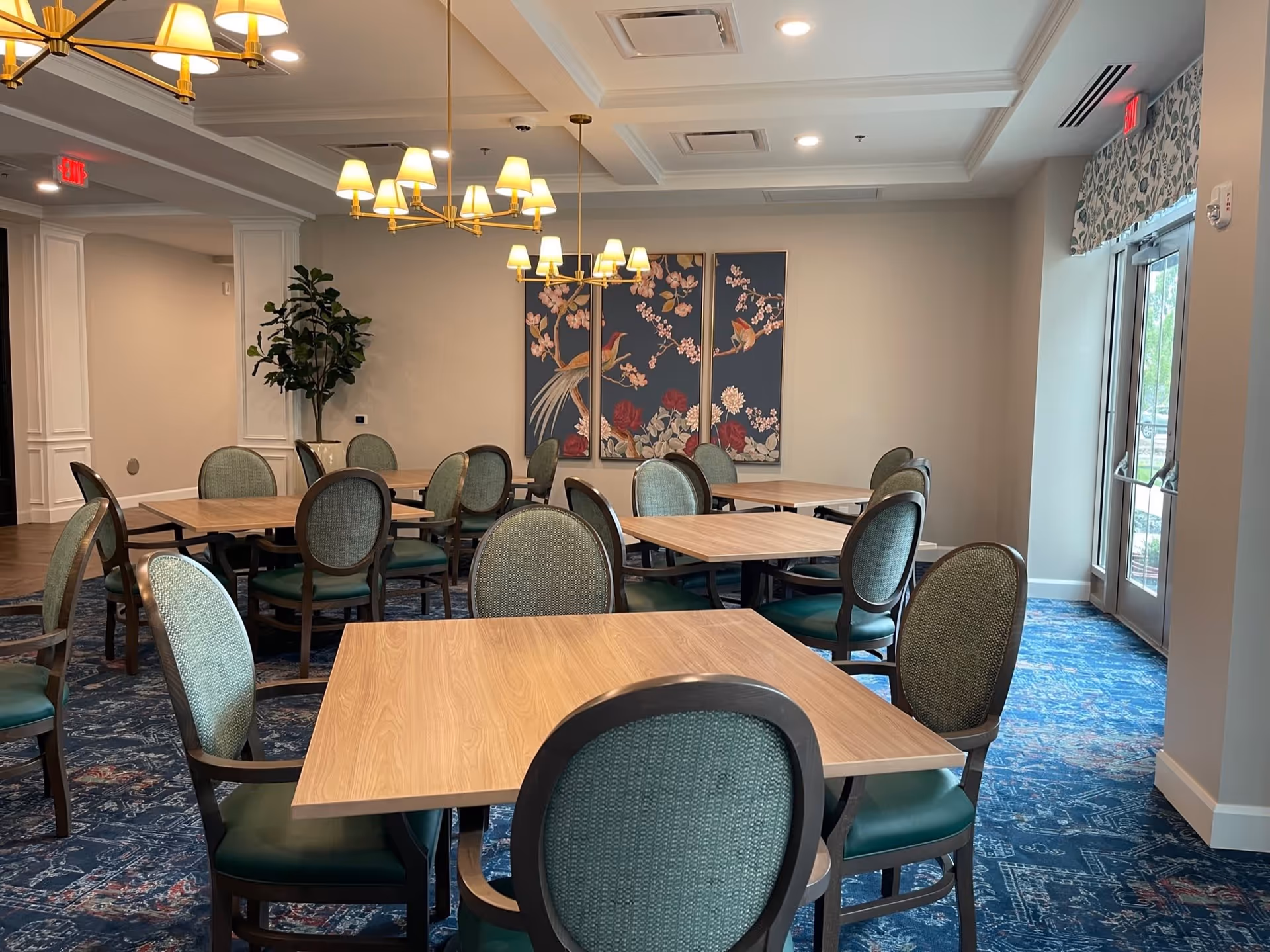 A dining room with multiple wooden tables and green cushioned chairs arranged neatly. The room features blue patterned carpet, two gold chandeliers with lampshades, a large plant in the corner, and a triptych wall art depicting birds and flowers. There is a glass door on the right side with a floral valance above it.