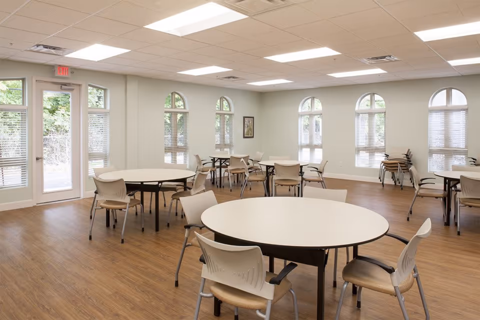 Bright common room with round tables and chairs, wood floors, arched windows, and an exit door.