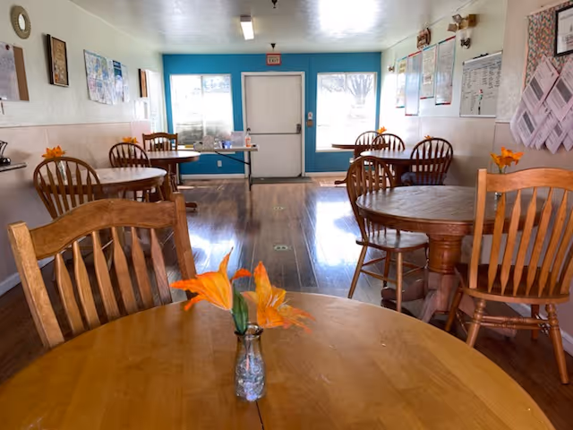 A bright dining room with wooden round tables and chairs. Each table has a small vase with orange flowers. The room has light-colored walls with various posters and notices pinned on them. There is a door and two windows at the far end letting in natural light.