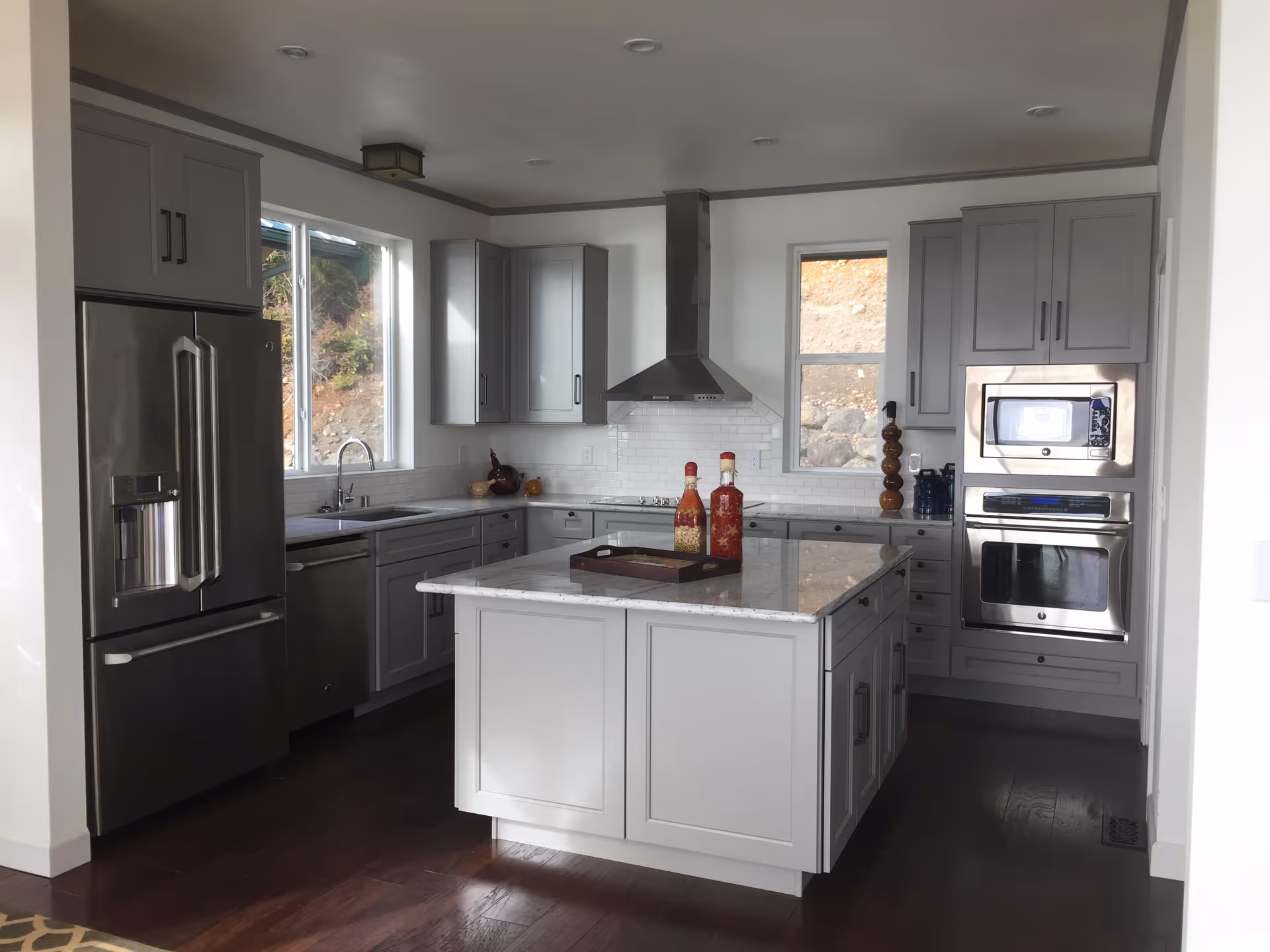 Modern kitchen with gray cabinets, stainless steel refrigerator, oven, microwave, and a central island with a marble countertop. Two decorative bottles are placed on the island. There are two windows letting in natural light and a stainless steel range hood above the stove.