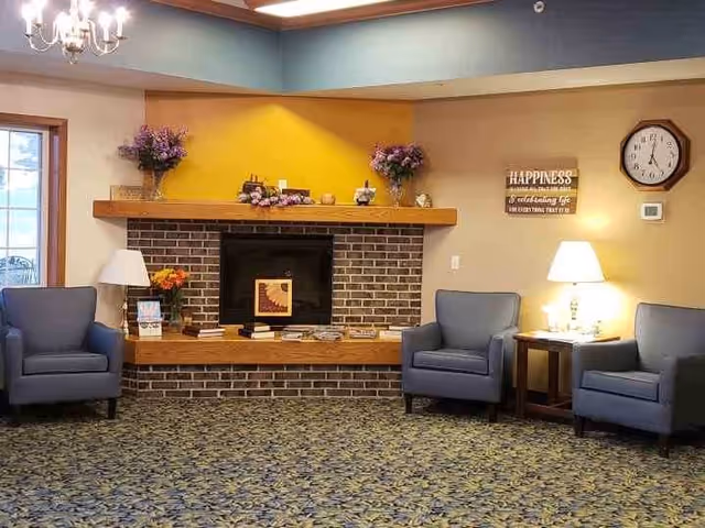 Cozy senior living common room with a brick fireplace, three blue armchairs, side tables with lamps, and decorative mantle items.