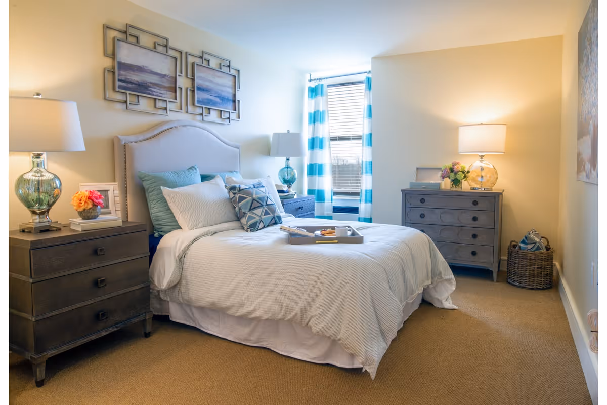 A cozy bedroom with a neatly made bed featuring white and blue pillows and bedding. There are two bedside tables with lamps and decorative items, a window with blue and white striped curtains, and a dresser with a lamp and flowers. The room has beige walls and carpeted flooring.