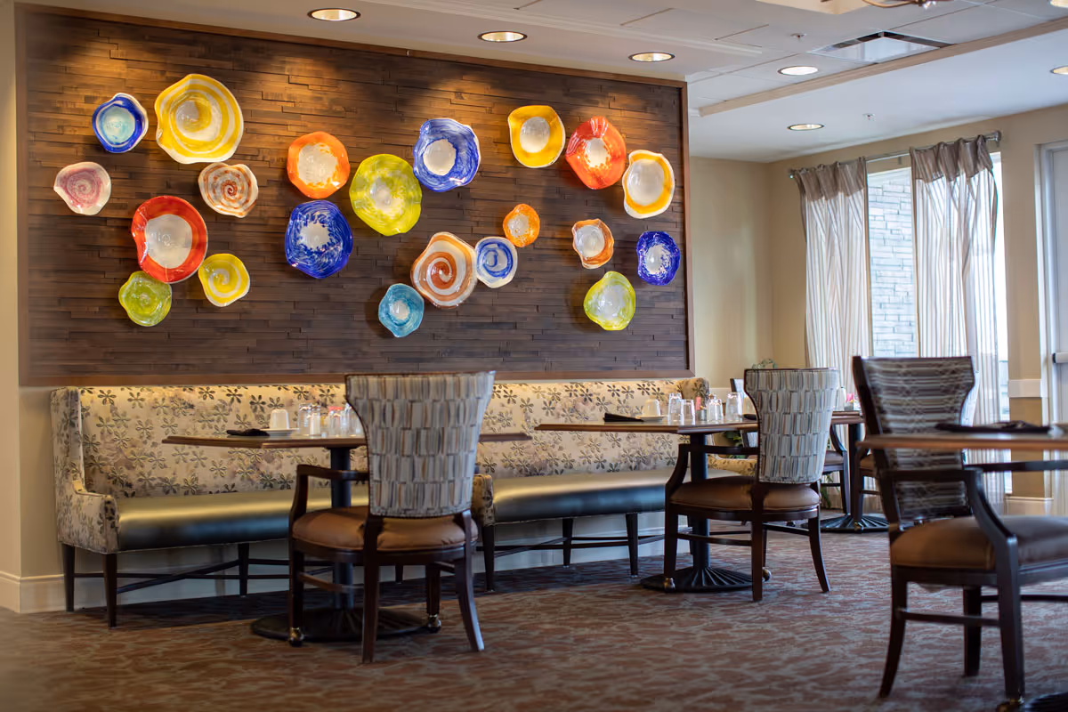 A dining area with tables and chairs arranged near a wall decorated with colorful, abstract ceramic plates mounted on a wooden panel. The space has large windows with sheer curtains allowing natural light to enter.
