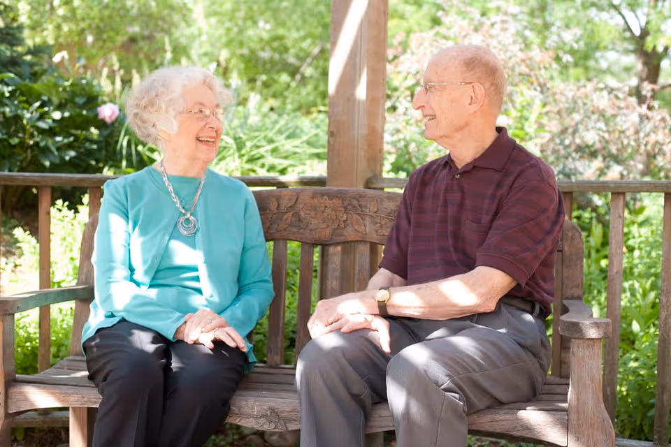 An elderly woman and an elderly man sitting on a wooden bench outdoors, smiling and engaged in conversation. The background shows green foliage and flowers, indicating a garden or patio setting.