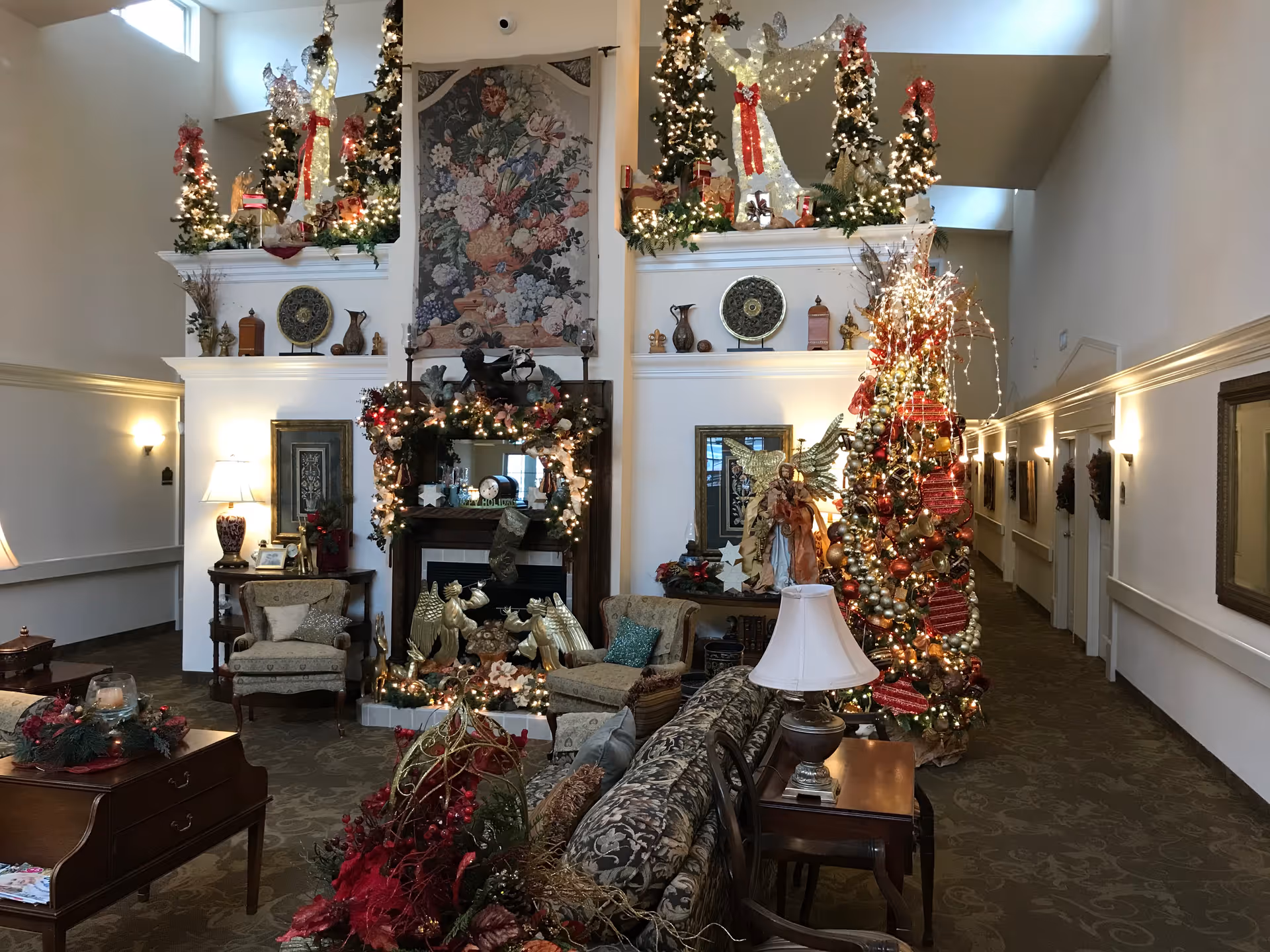 A warmly decorated living room area in Dudneywood Assisted Living, featuring a Christmas tree adorned with red and gold ornaments and lights. The room includes a fireplace decorated with garlands and stockings, two armchairs, a patterned sofa, side tables with lamps, and various holiday decorations including angels and wreaths. The walls are light-colored with framed artwork and sconces, and there is a hallway visible to the right.