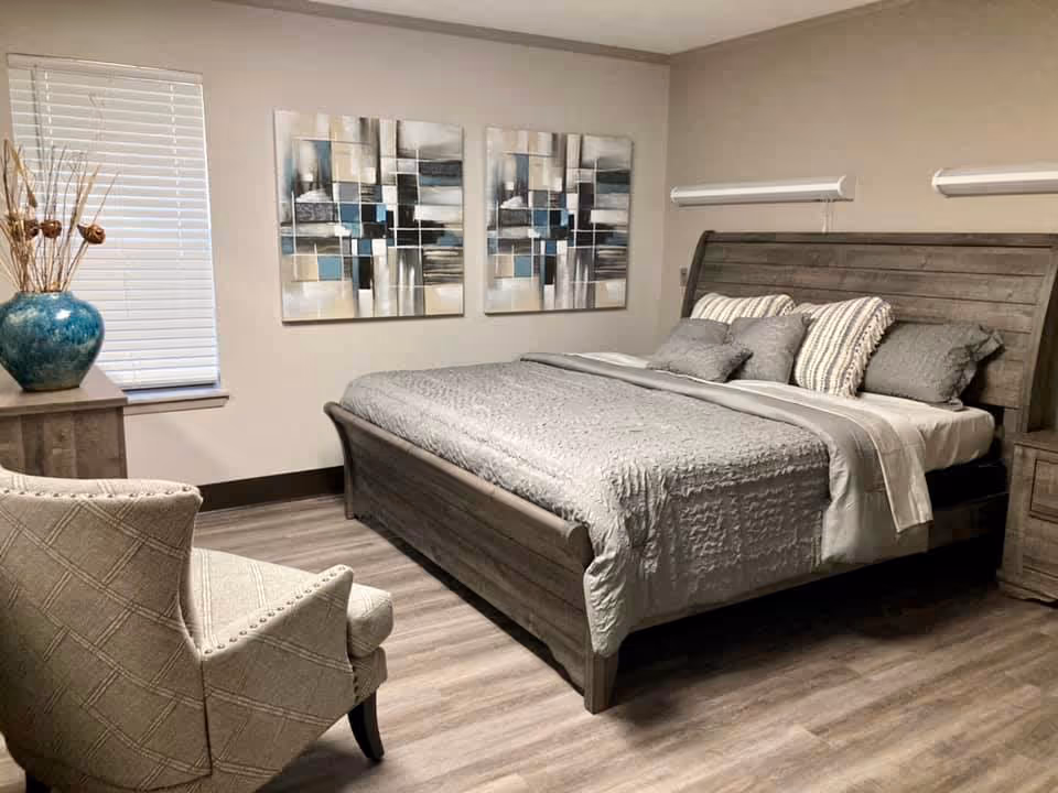 A neatly arranged bedroom with a large wooden bed featuring gray bedding and multiple pillows. There is a cushioned armchair with a diamond pattern on the left side, a wooden dresser with a large blue vase holding decorative branches, and two abstract paintings with blue, beige, and black tones hanging on the wall above the bed. The room has light-colored walls and wood flooring.