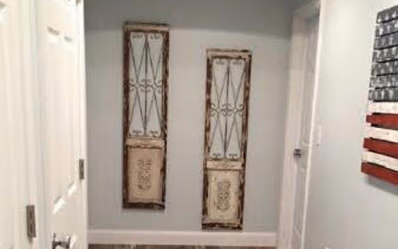 A hallway with light gray walls and white doors on either side. Two decorative wall hangings with rustic wooden frames and metal designs are mounted on the wall at the end of the hallway. A wooden American flag decoration is partially visible on the right wall.