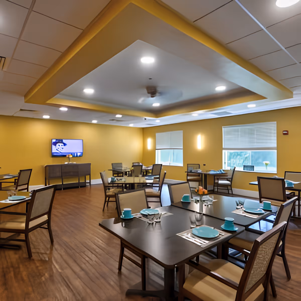 A dining room with multiple tables and chairs arranged neatly. Each table is set with turquoise plates, cups, and silverware on placemats. The room has yellow walls, wooden flooring, and a ceiling with recessed lighting and a ceiling fan. A TV is mounted on one wall above a sideboard, and windows with blinds allow natural light into the space.