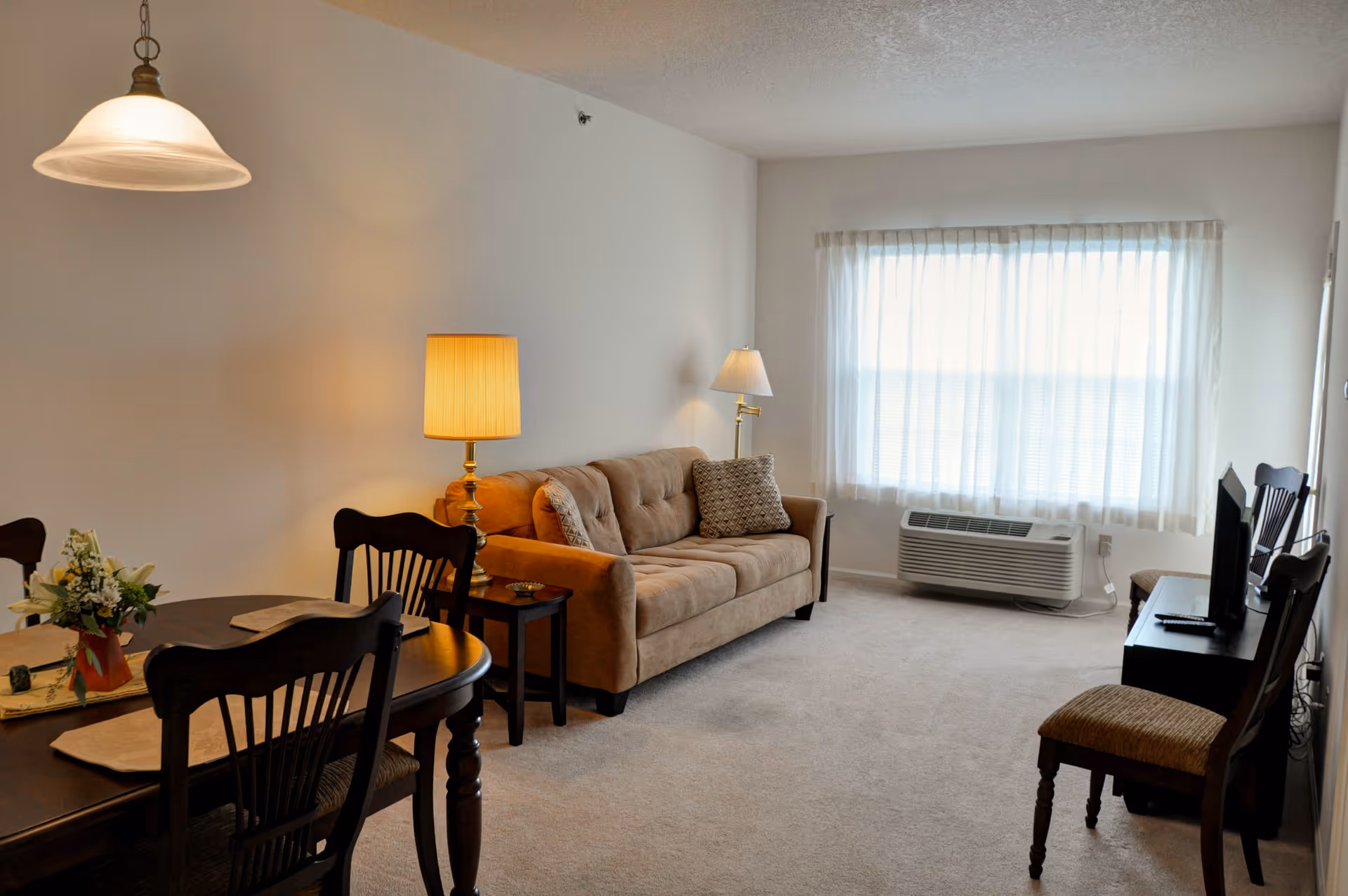 A cozy living room with a beige sofa, two side tables with lamps, a window with sheer curtains, a small TV on a stand, and a dining table with chairs and a flower arrangement.