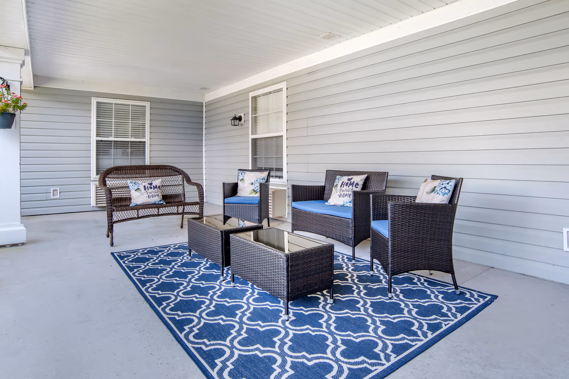 Covered outdoor patio area with gray siding walls, featuring a blue and white patterned rug, dark wicker seating including a loveseat, two armchairs, and a bench with cushions and decorative pillows that say 'Home Sweet Home'.