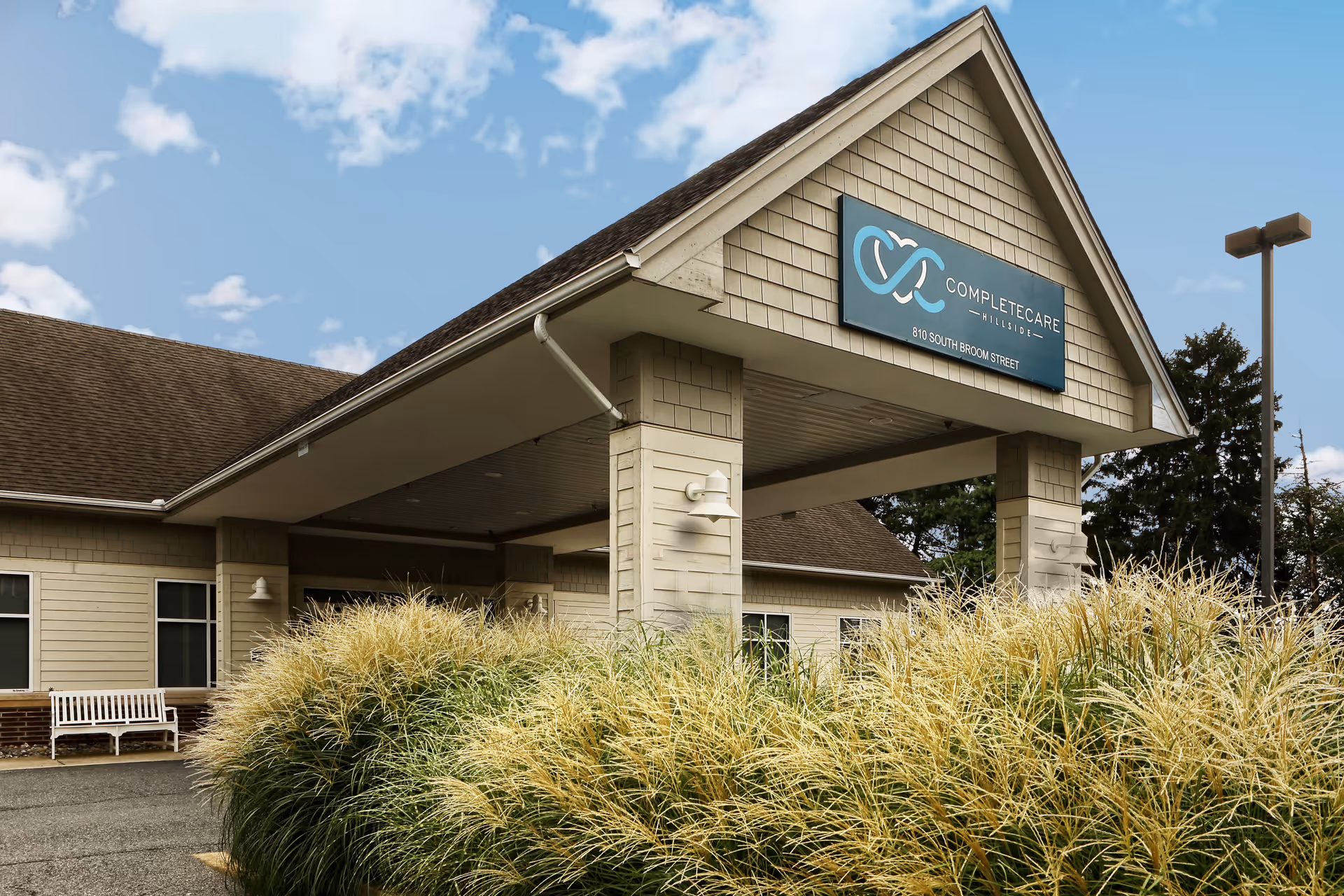 Exterior view of the entrance to Complete Care at Hillside facility with a covered drop-off area, beige siding, a sign displaying the facility name and address, ornamental grasses in the foreground, and a partly cloudy sky.