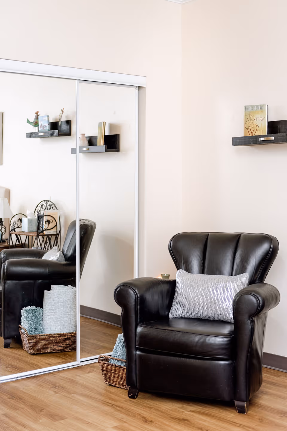 A cozy corner of a room featuring a black leather armchair with a silver decorative pillow. Behind the chair is a wall with a small floating shelf holding a book titled 'The Mystery of God's Will.' To the left, there is a large mirrored sliding closet door reflecting part of the room, including a small table with a lamp and decorative items. A woven basket with folded blankets is placed on the wooden floor next to the chair.