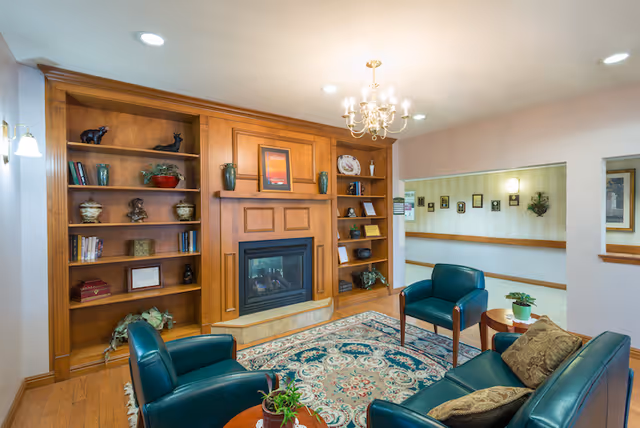 A cozy common room with built-in wooden bookshelves and a fireplace, teal leather chairs, a sofa, and a patterned rug under a chandelier.