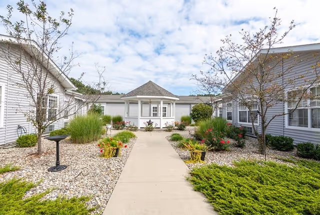 A landscaped courtyard with a concrete walkway leading to a small gazebo structure. The courtyard is surrounded by single-story buildings with white siding and multiple windows. There are various plants, shrubs, and small trees planted in gravel beds on either side of the walkway under a partly cloudy sky.