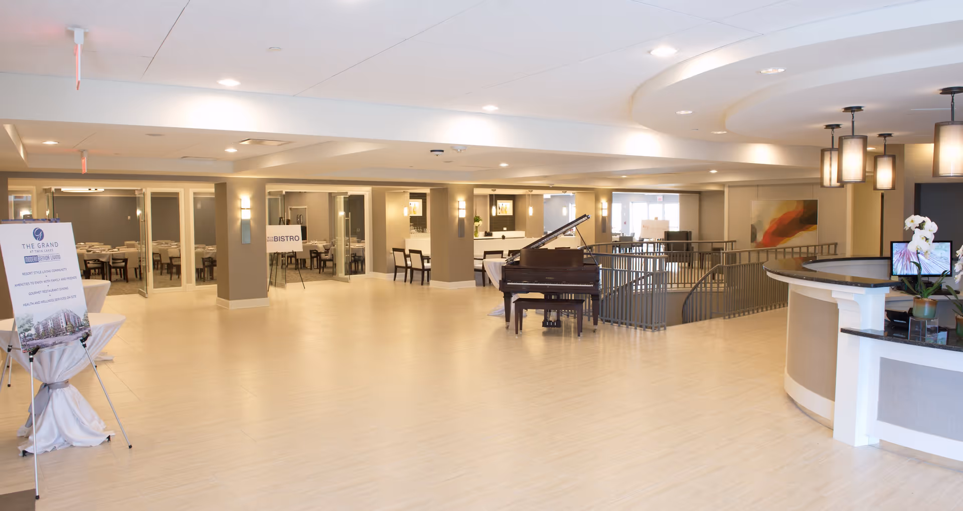 Spacious, well-lit interior of a senior living facility lobby area with light-colored flooring and walls. A grand piano is positioned near a railing that overlooks a lower level. To the left, there is a sign on an easel and a dining area labeled 'Bistro' visible through glass doors. On the right, there is a curved reception desk with a computer and a small plant. Modern ceiling lights illuminate the space.