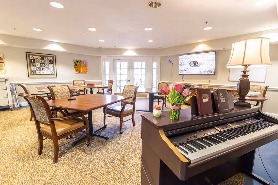 A bright and spacious common area with several wooden tables and cushioned chairs arranged on a patterned carpet. A digital piano with sheet music and a vase of pink tulips is in the foreground. The room has light-colored walls adorned with framed artwork, a flat-screen TV mounted on the wall, and large glass doors letting in natural light.