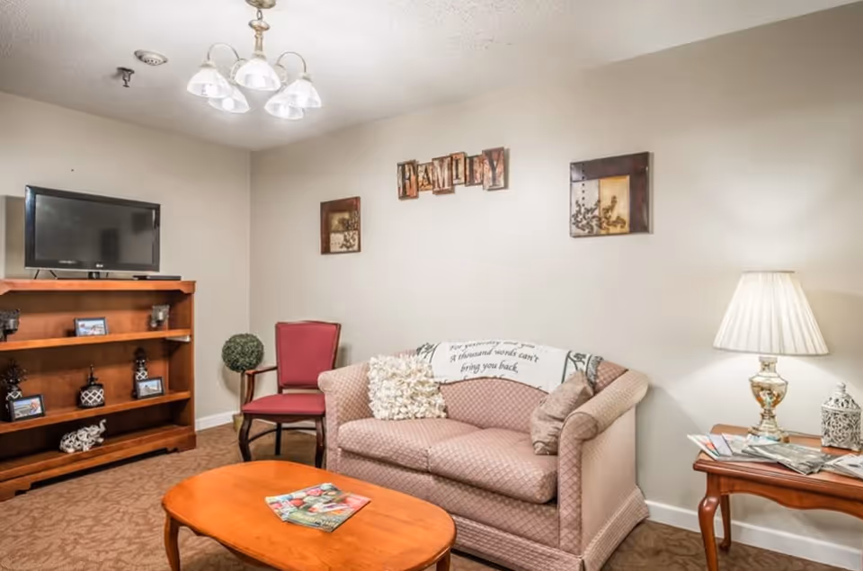 A cozy living room with a patterned pink loveseat, a red armchair, a wooden coffee table with magazines, and a wooden TV stand holding a flat-screen television and decorative items. The walls are decorated with framed art and a sign that reads 'FAMILY'. A table lamp sits on a side table next to the loveseat.