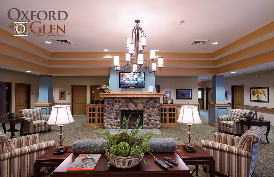 Bright memory care lounge with a stone fireplace, TV, chandelier, and striped armchairs arranged around coffee tables.