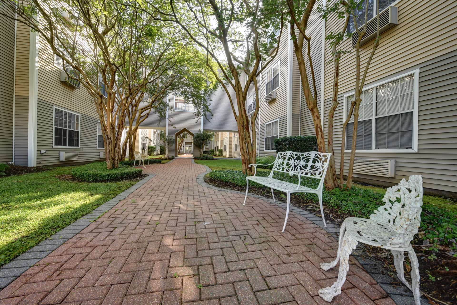Brick-paved courtyard walkway between senior living buildings lined with trees and white decorative benches.