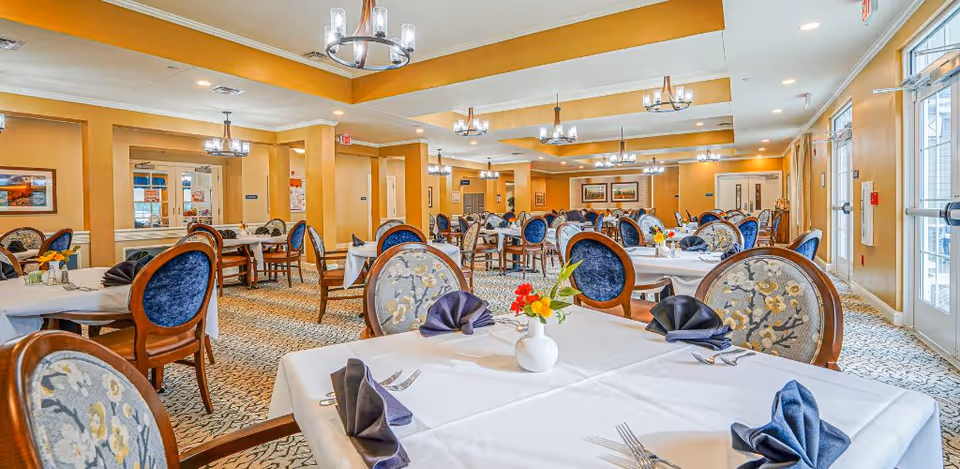 A spacious dining room with multiple round tables covered in white tablecloths, each set with folded navy blue napkins, silverware, and small floral centerpieces. The room features upholstered chairs with blue and floral patterned backs, warm yellow walls, carpeted floors, and modern chandeliers hanging from the ceiling. Large windows on the right side allow natural light to brighten the space.