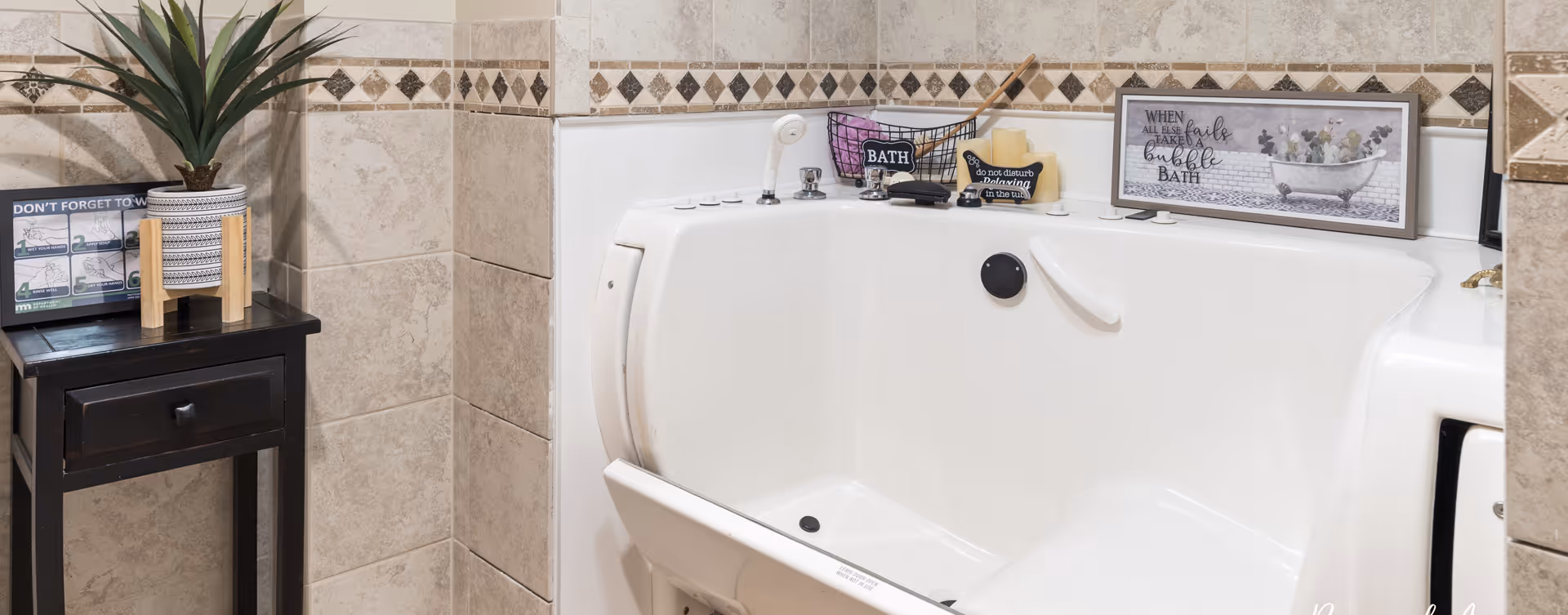 A walk-in bathtub with a door for easy access, surrounded by beige tiled walls with decorative trim. On the edge of the tub are bath items including soap and a small basket, along with a framed sign that reads 'When all else fails take a bubble bath.' To the left of the tub is a small black wooden table with a potted plant and a sign reminding to not forget to wash hands.