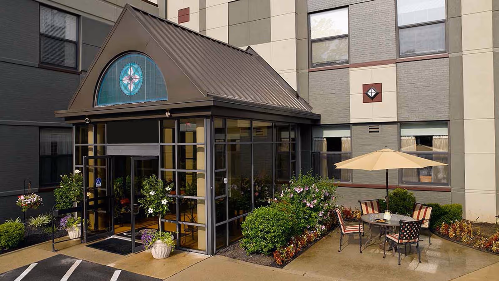 Exterior entrance of a senior living facility with a glass-enclosed entryway featuring a decorative stained glass window above the door. There are potted plants and bushes around the entrance, and a patio area with a round table, four chairs with striped cushions, and a large beige umbrella. The building has multiple windows and a beige and gray color scheme.
