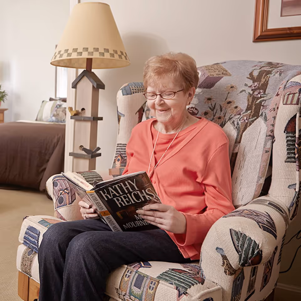 An elderly woman wearing glasses and a coral sweater is sitting comfortably in a patterned armchair, reading a book by Kathy Reichs titled 'My Mourning'. Behind her is a lamp with a birdhouse design and a bed with pillows in the background.