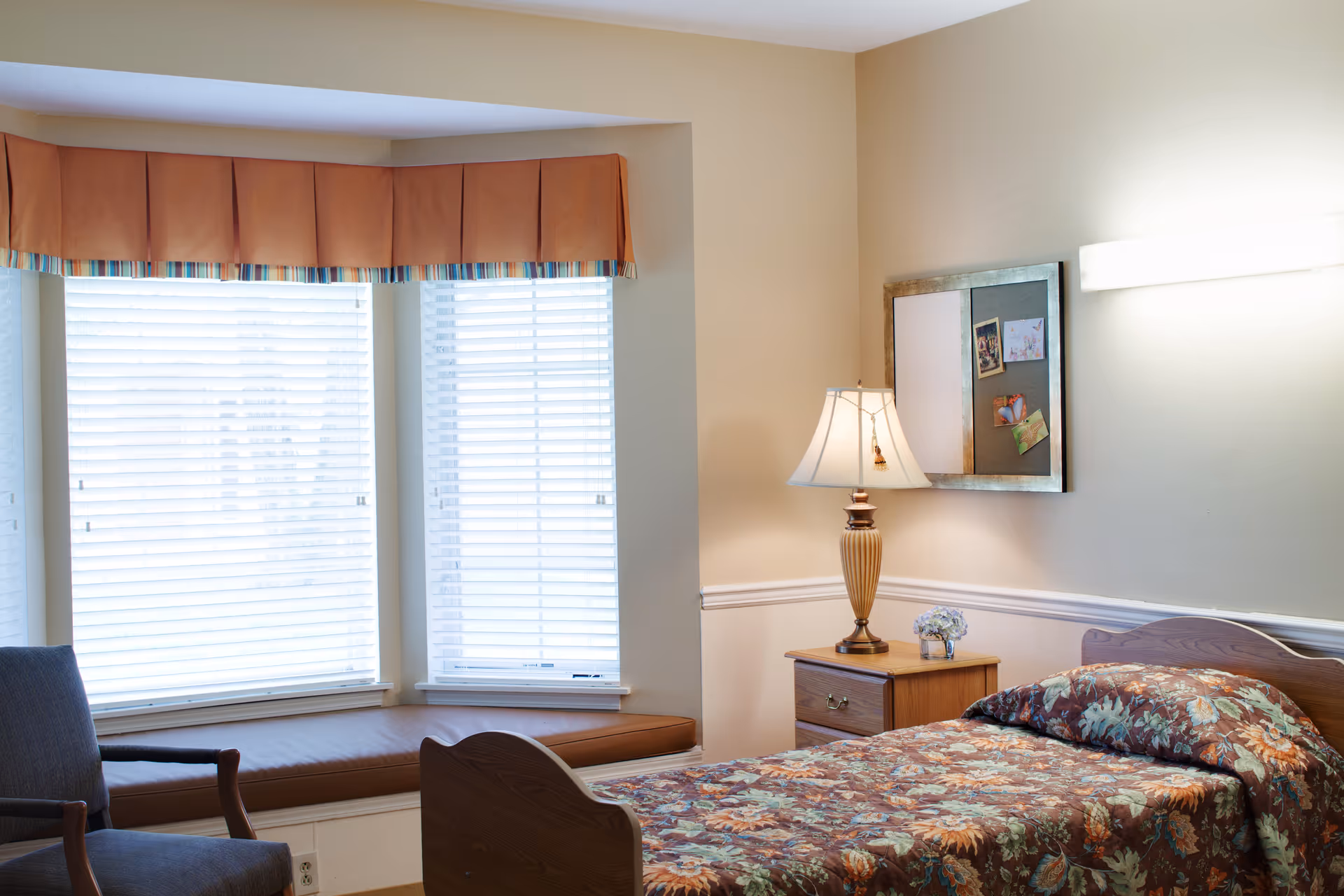 Sunlit bedroom with a single floral bed, wooden nightstand and lamp beside a bay window with a cushioned window seat.