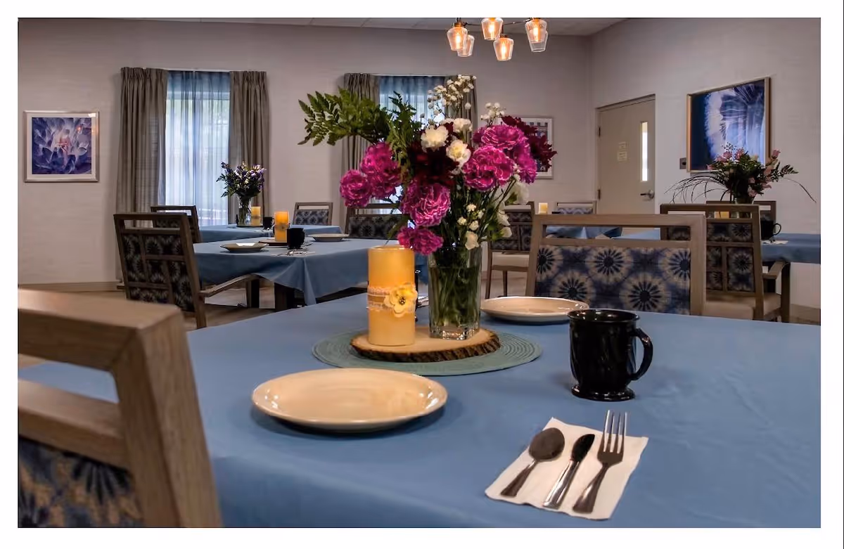 A dining room with tables covered in blue tablecloths, each set with plates, utensils, and mugs. A vase with vibrant pink and white flowers and a lit candle are placed on the nearest table. The room has several chairs with patterned cushions, framed artwork on the walls, and windows with curtains allowing natural light.