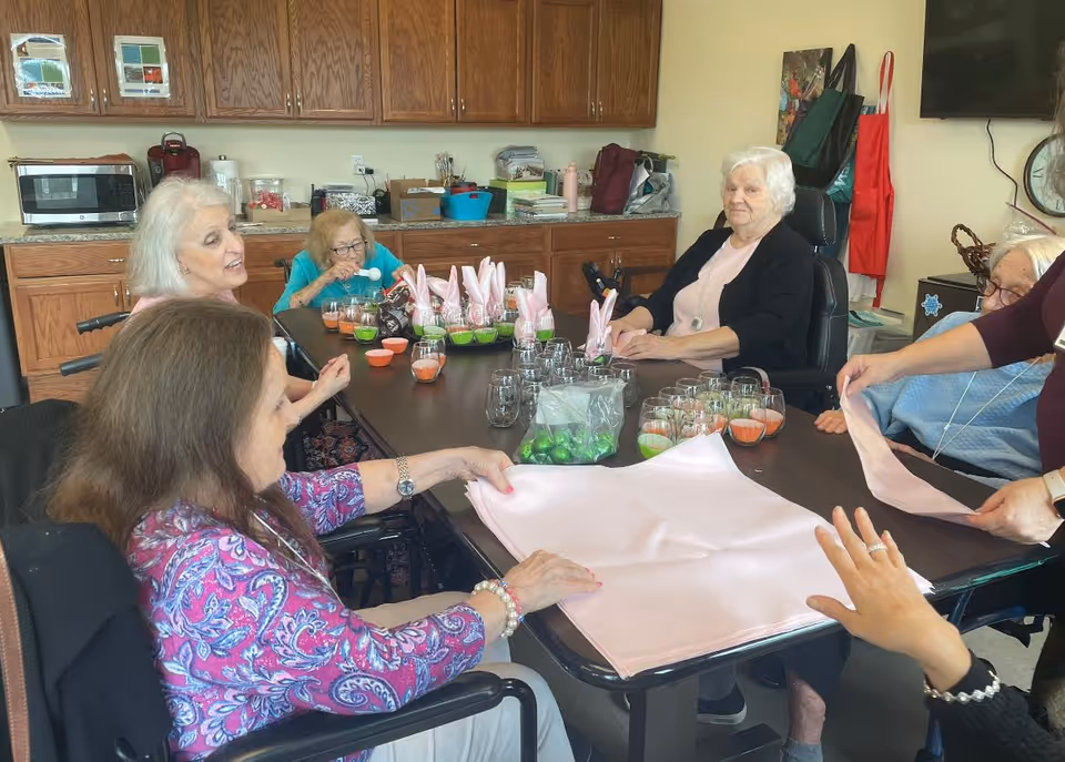 A group of elderly women sitting around a table in a kitchen or dining area, engaging in an activity involving pink napkins and small cups with colorful contents. The room has wooden cabinets, a microwave, and various items on the countertop.